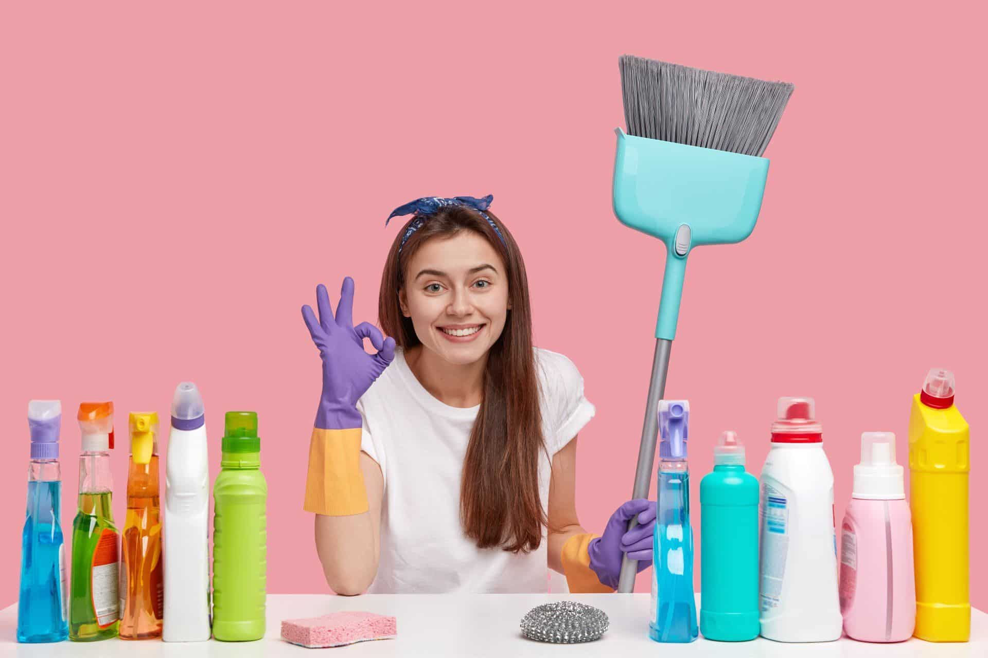 a woman with cleaning supplies and holding a broom making the 'okay' sign with her hand
