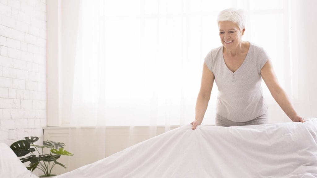 An elderly woman smiling and making her bed as as tep for cleaning a room in 5 minutes.