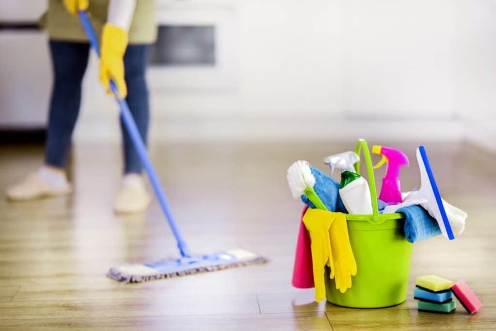 A green bucket filled with house cleaning tools as someone mops in the background.
