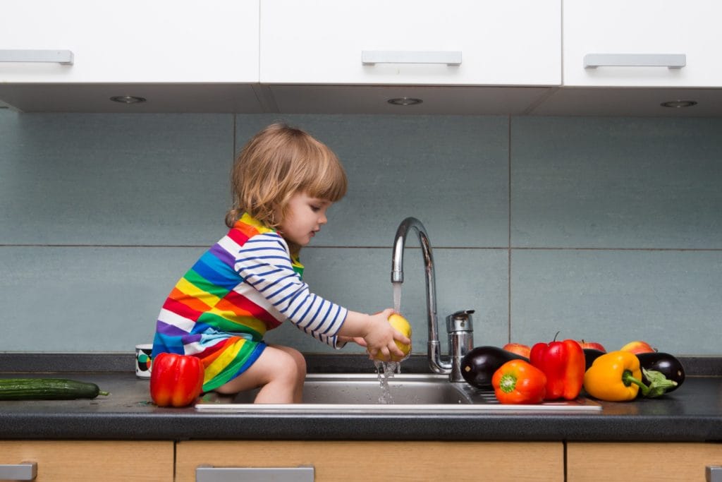 A toddler sitting on a counter washing produce.