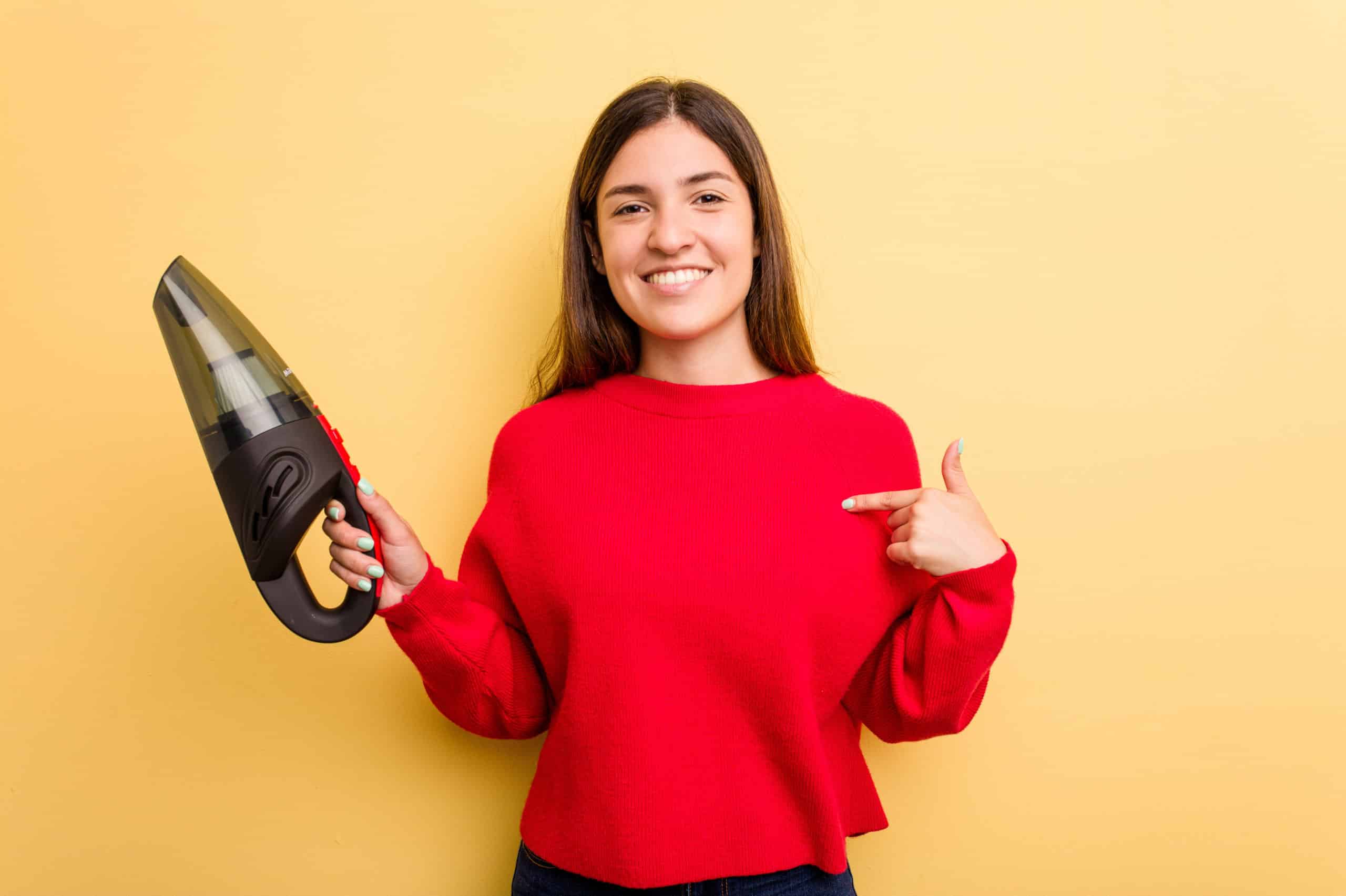 Young caucasian woman holding a hand vacuum cleaner isolated on yellow background person pointing by hand to a shirt copy space, proud and confident