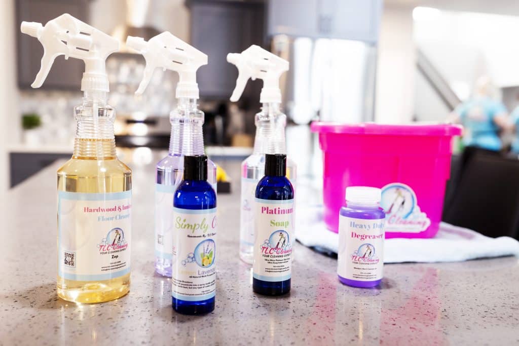 A variety of labeled cleaning products are lined up on a speckled countertop, including sprays and a degreaser, with a pink cleaning bucket in the soft-focused background.