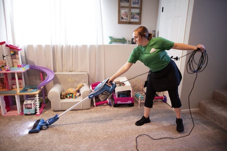 A professional cleaner providing deep cleaning services in a home, using a handheld vacuum cleaner to tidy a carpeted living area cluttered with children's toys.