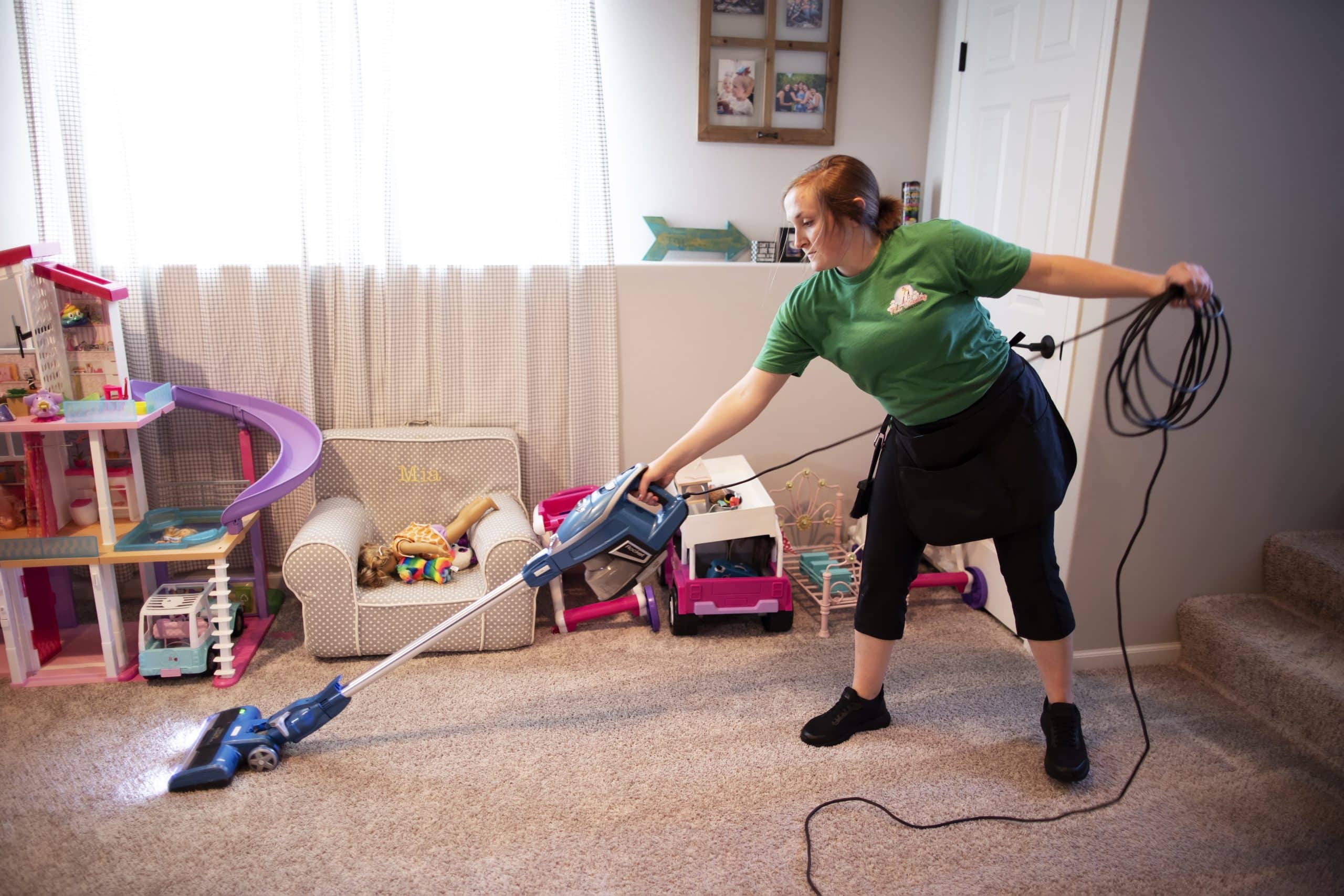 A professional cleaner providing deep cleaning services in a home, using a handheld vacuum cleaner to tidy a carpeted living area cluttered with children's toys.
