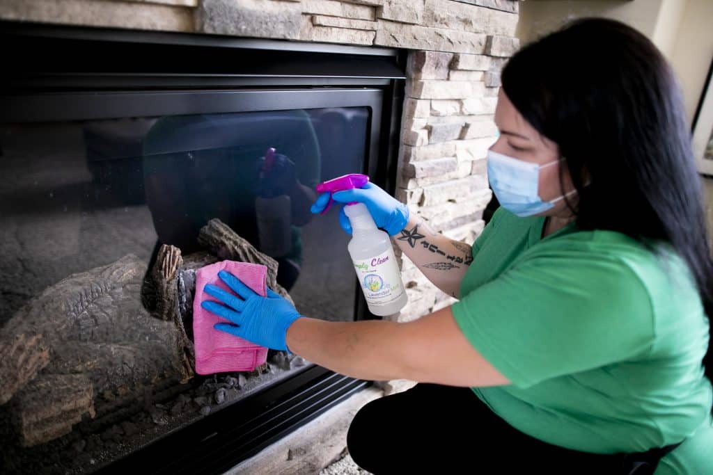 A person wearing a mask and blue gloves provides deep cleaning services, meticulously wiping down a fireplace glass door with a pink cloth and spray cleaner.
