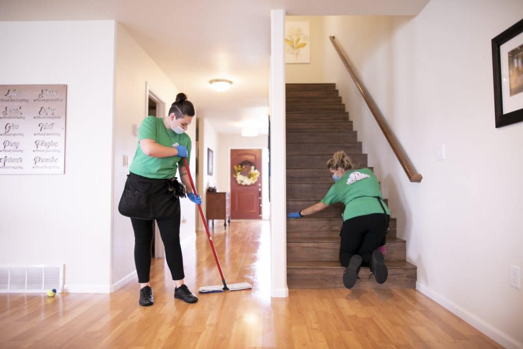 Two cleaning professionals in green shirts performing a thorough cleaning: one mopping the hardwood floor, and the other scrubbing the stairs in a well-lit home entryway.