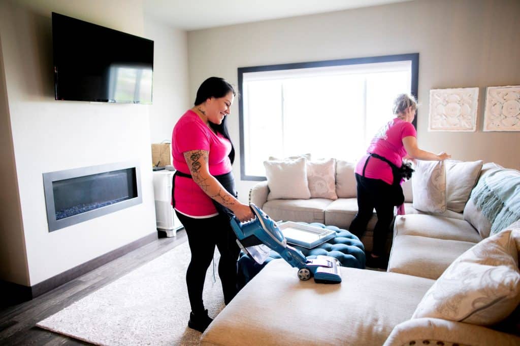 Two professional cleaners in pink shirts perform a thorough cleaning in a living room, vacuuming the carpet and dusting the furniture.