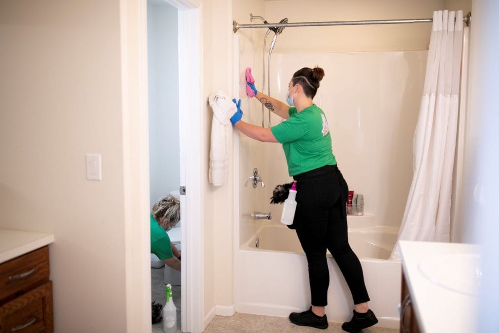 Two professional cleaners in green uniforms clean a bathroom, with one scrubbing the shower wall and the other cleaning the toilet in the adjacent room.